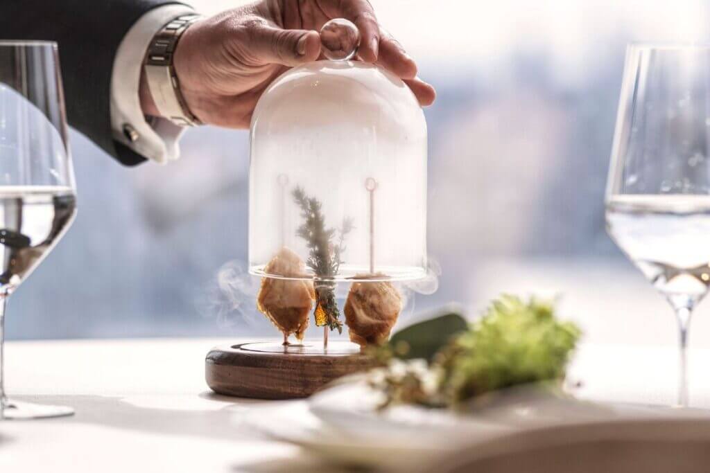 A waiter lifts the cloche on a dish of crispy smoked chicken wing, lemon and spices 