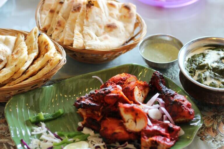 Tandoori chicken in a banana leaf bowl with two baskets of naan, a small metal bowl of green-coloured raita, and a bigger metal bowl of a white and green curry, served at an Indian Restaurant.
