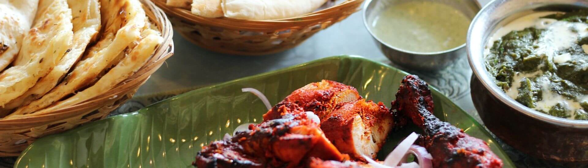 Tandoori chicken in a banana leaf bowl with two baskets of naan, a small metal bowl of green-coloured raita, and a bigger metal bowl of a white and green curry, served at an Indian Restaurant.